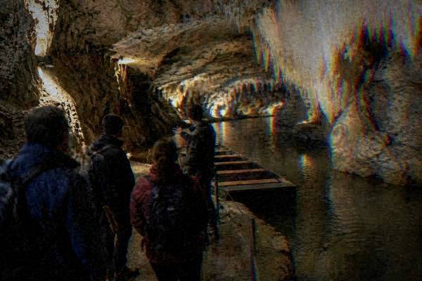 A guide and a small group at the wet stone mouth of a lit limestone cave with smooth pale stalactites, soft warm lamps, Punkva Caves
