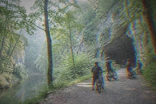 A small group on e-bikes with helmets stopped on a gravel track at the wooded mouth of a limestone cave in a green gorge, Sloup-Šošůvka Caves