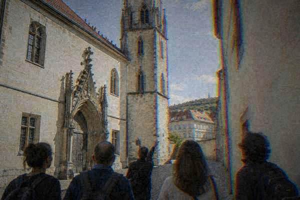 A guide pointing up at a slender Gothic stone tower while a small group looks on from a narrow cobbled passage, Old Town Hall