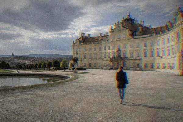 A lone visitor seen from behind on a broad gravel forecourt before a huge Baroque chateau with a long pale curved front, Valtice Chateau