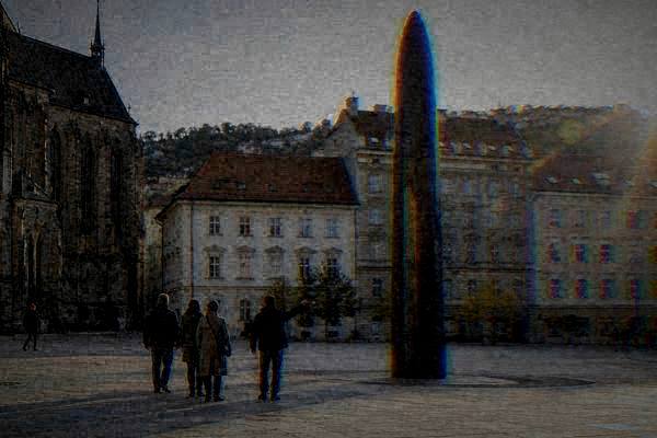 A guide and a small group seen from far behind on a wide pale paved square beside a tall slim column of dark polished stone, Brno Astronomical Clock