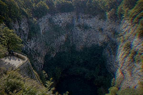 A few visitors small in the frame on a stone viewing platform on the rim of a huge round limestone sinkhole ringed by beech forest, Macocha Abyss