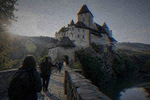 A guide and a few visitors crossing a stone bridge toward a vast medieval castle of many stacked towers on a rocky spur, Pernštejn Castle