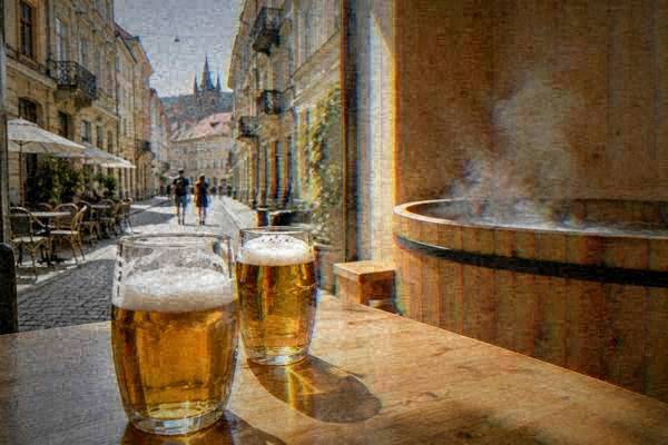 Two glasses of golden beer on a wooden table in a bright room with pale timber walls, steam rising from a round wooden tub behind, no faces in the frame, Kobližná Street