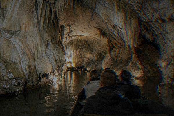 A few visitors seen from behind on a flat-bottomed boat gliding on a dark underground river between pale wet limestone walls lit by soft warm lamps, Punkva Caves