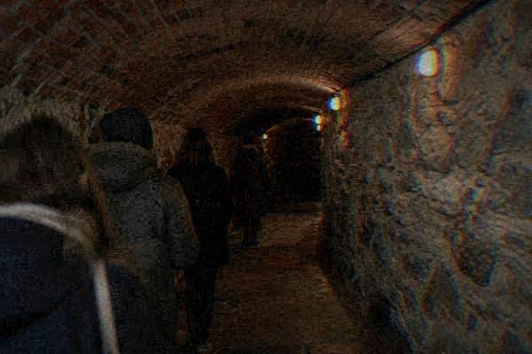 A guide and a few visitors seen from behind in a low brick-vaulted passage with rough stone walls and warm low lamps, Labyrinth under the Cabbage Market