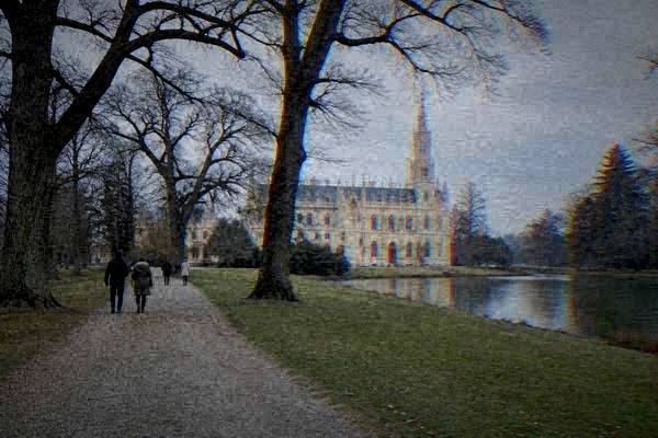 A small group walking a gravel avenue between old oaks toward a pale neo-Gothic chateau with a tall slender tower, Lednice and Valtice cultural landscape