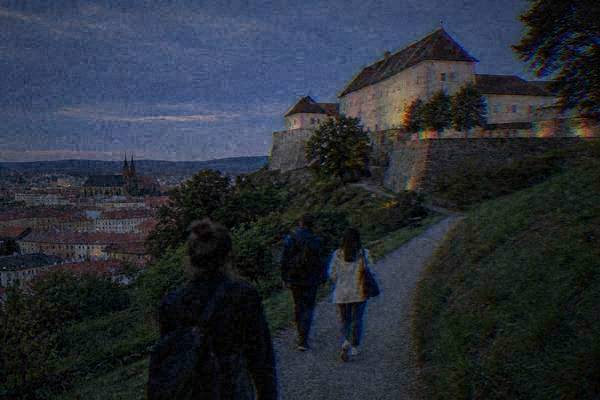A private guide and two visitors climbing a gravel path through green slopes toward a hilltop fortress with pale walls, Spilberk Castle