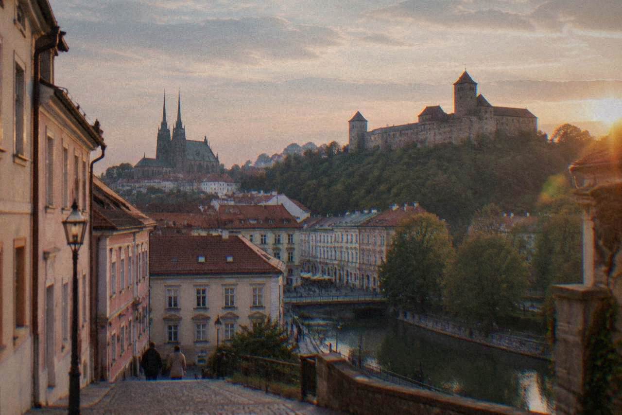 A stone castle fortress with high walls and towers standing above the rooftops of Brno at dusk
