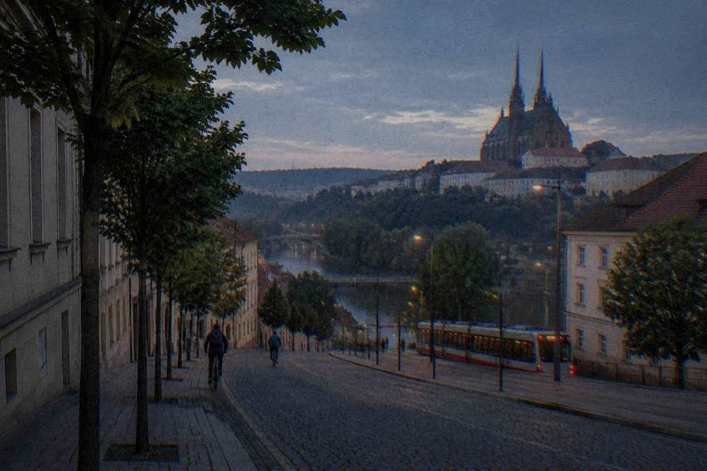 A tree-lined street of low older buildings sloping down toward a river in a Central European city