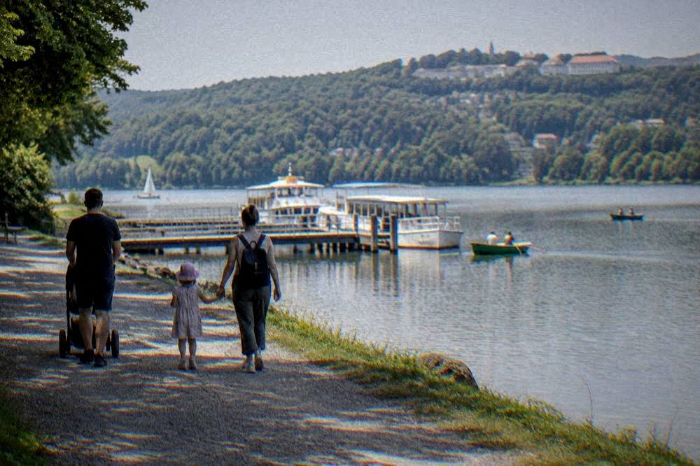 A family walking a gravel path along a lakeshore with wooded hills behind