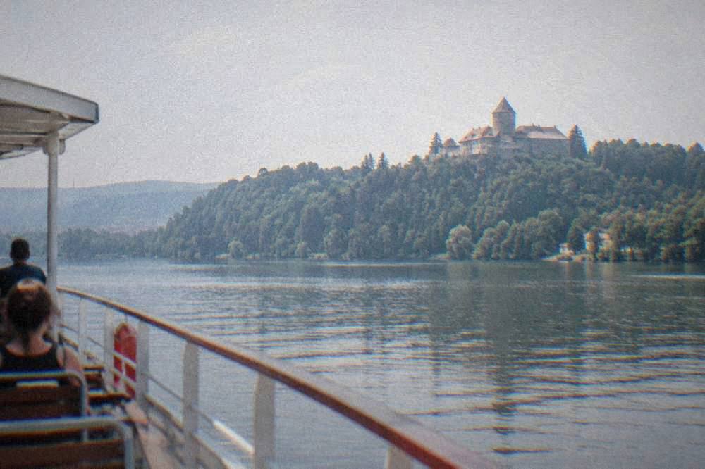 A stone castle on a forested hill seen across a wide stretch of water from a boat deck