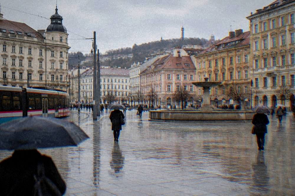 People with umbrellas crossing a wide square in light rain between historic buildings