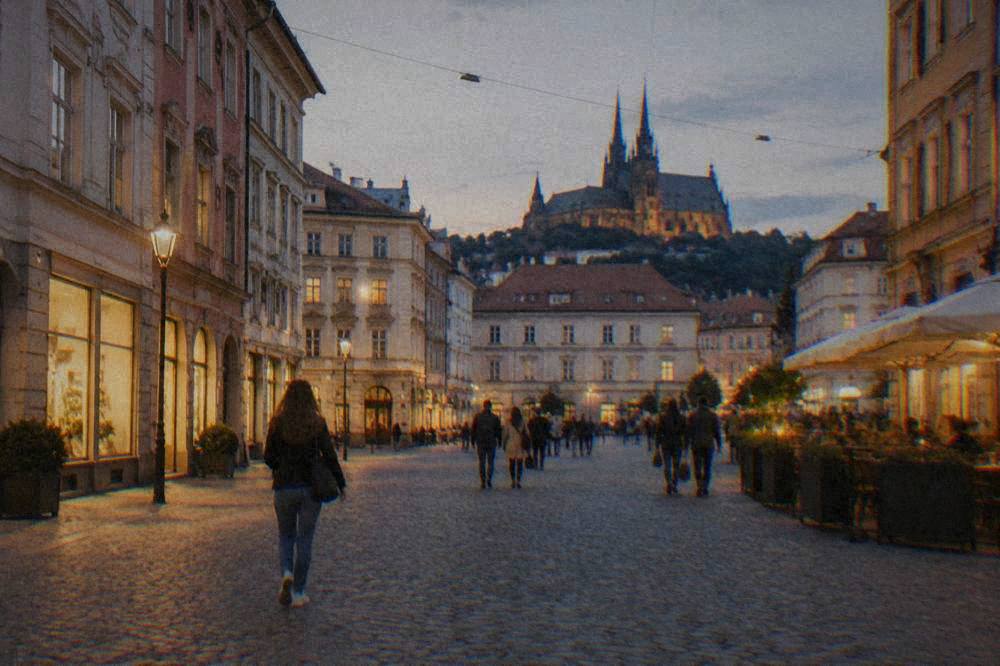 An evening view of a Brno square with lit shopfronts and people walking