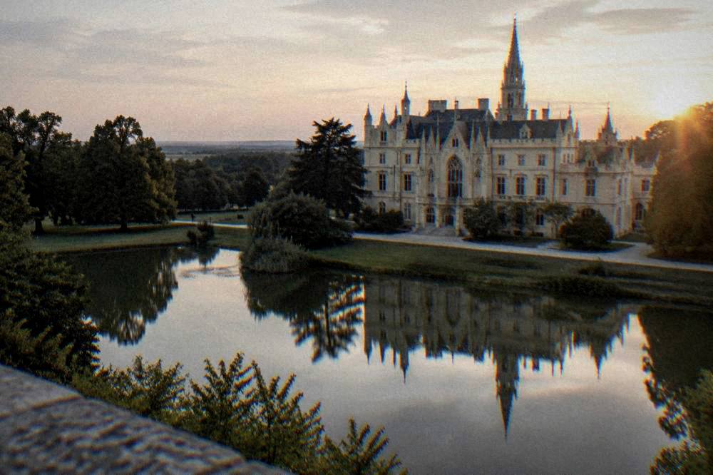 A stone chateau facade with ornate windows reflected in a still pond at dusk