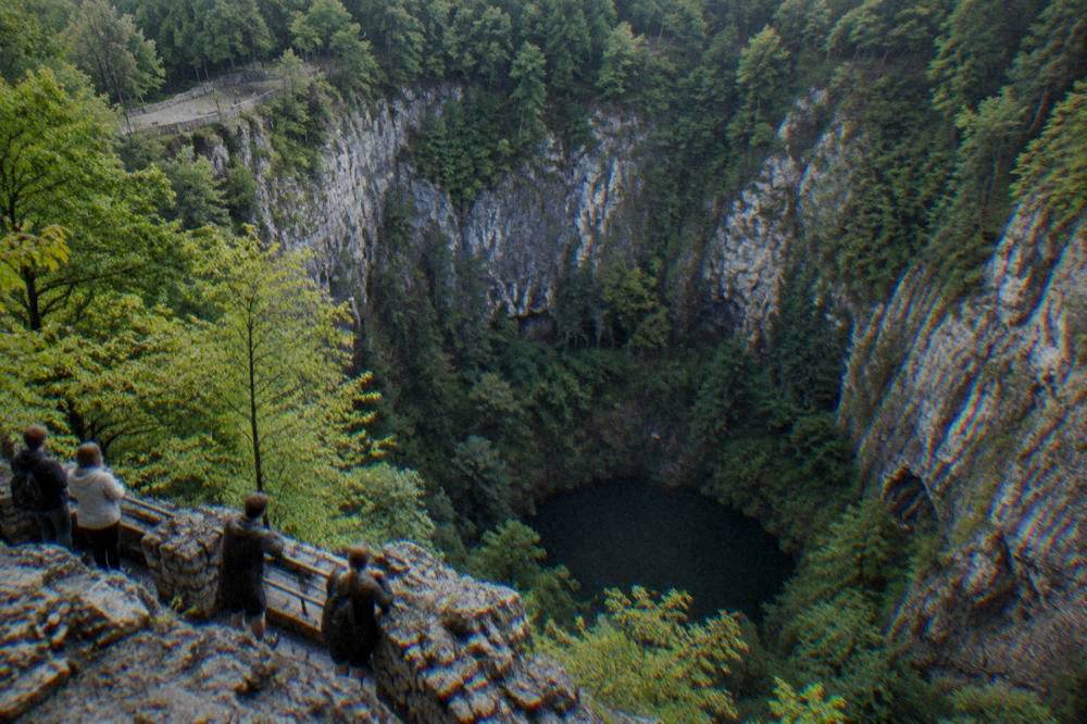 Visitors leaning on a railing at a rocky overlook above a deep green gorge