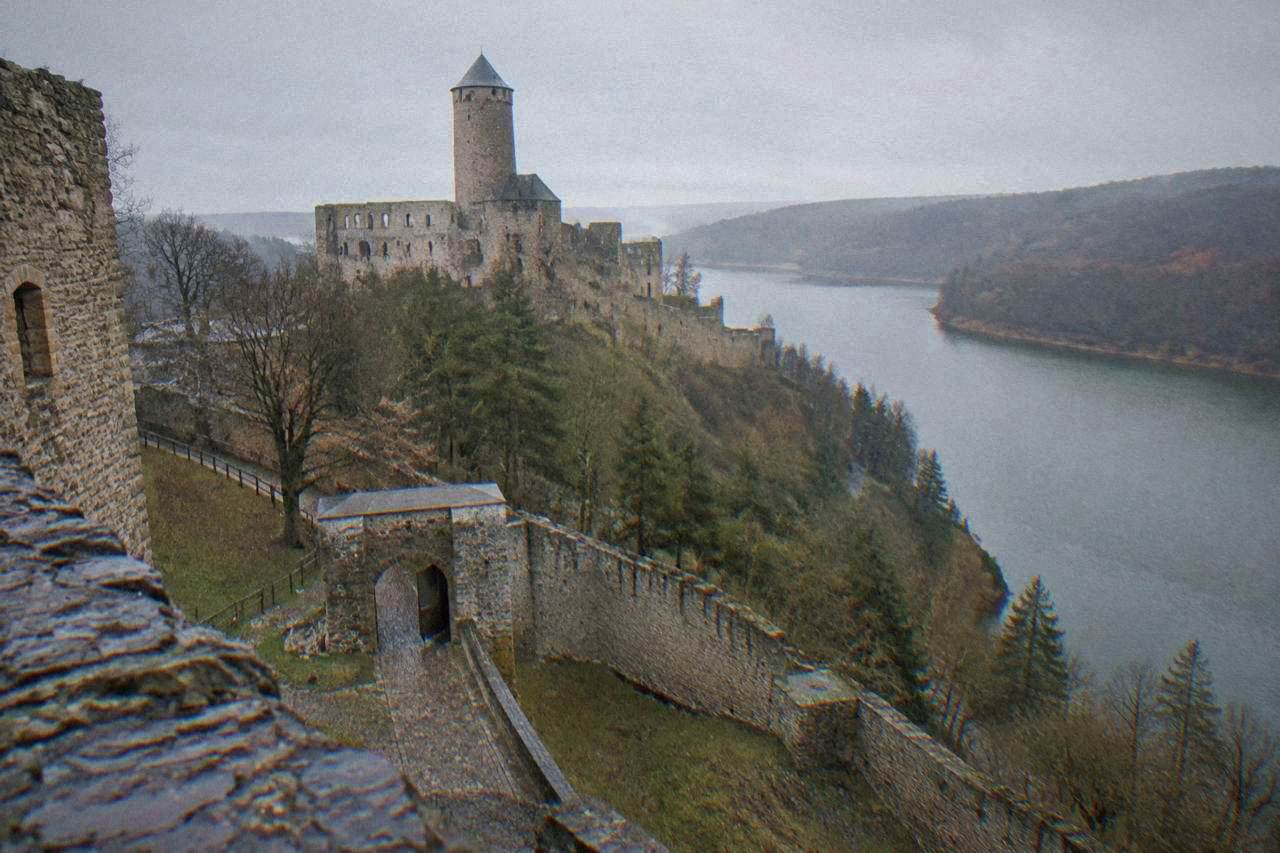 A stone castle with a tall round tower standing on a wooded promontory above a wide river reservoir