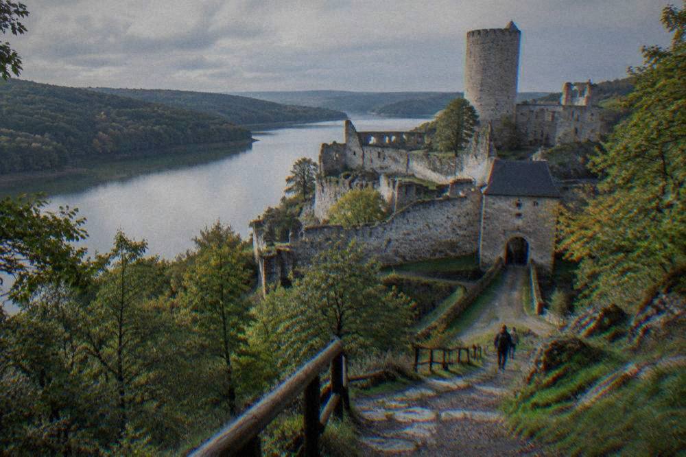 A forested hillside path leading up toward castle walls with views over water