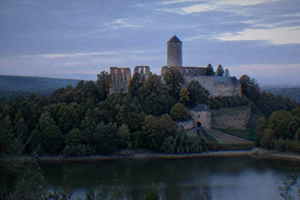 A large medieval castle on a forested ridge seen from across a valley