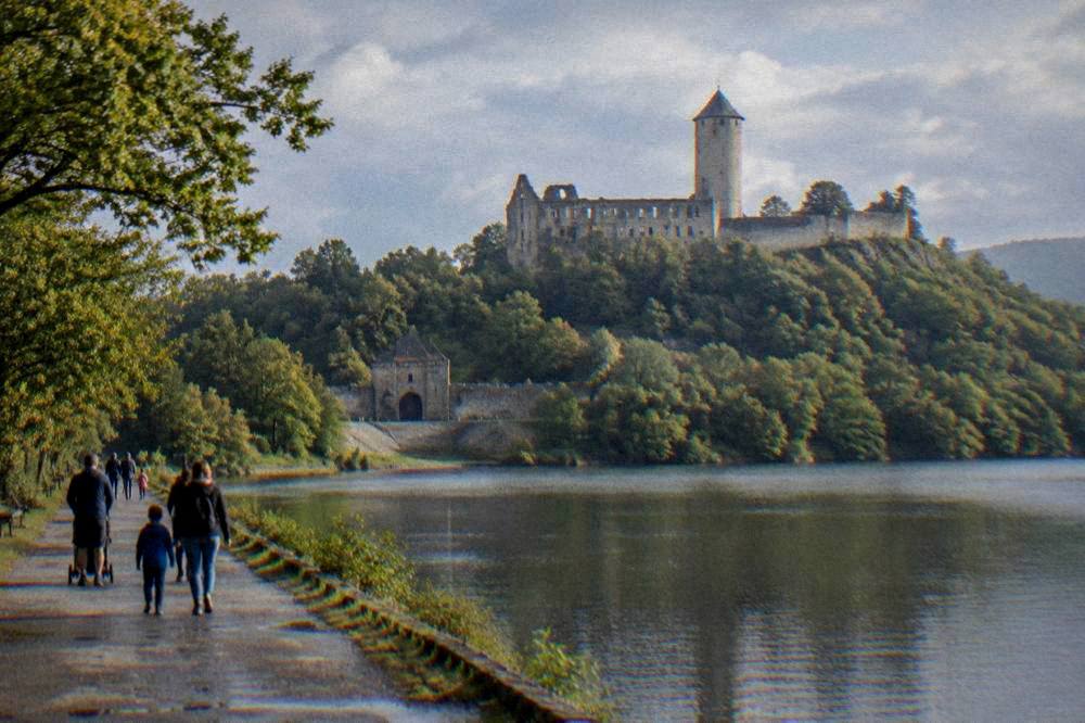 Families walking a lakeside promenade with a castle visible on a hill in the distance
