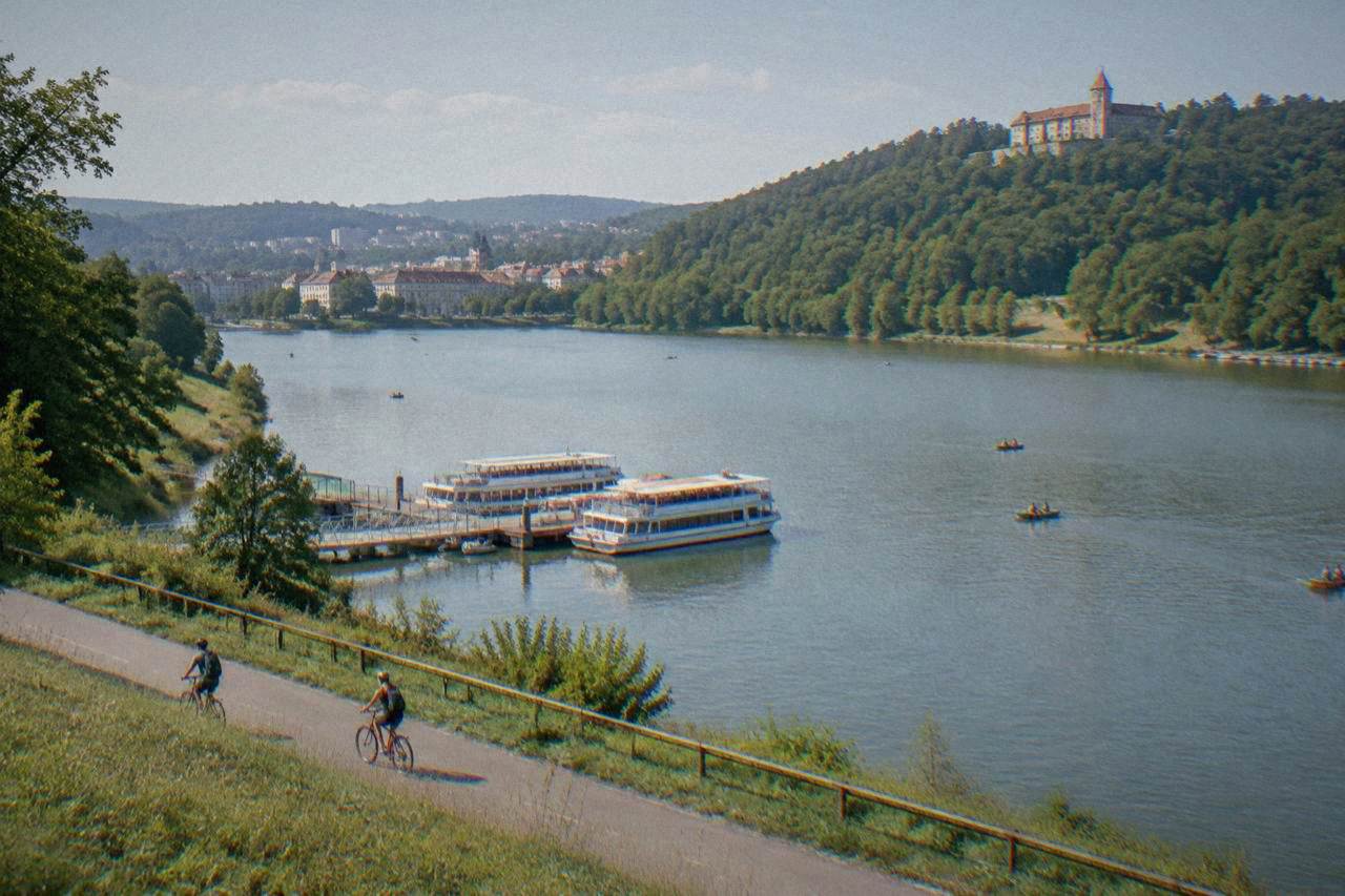 Cyclists riding along a riverside path with a forested hillside and a castle in the background