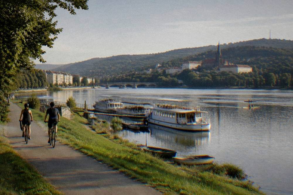 Two cyclists on a riverside bike path in evening light with a stone bridge crossing the river
