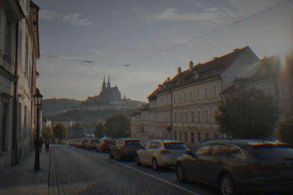 A row of cars parked along a street in Brno with striped parking bays and old buildings behind them
