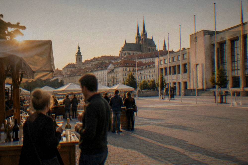 People tasting wine at outdoor stalls on a sunny historic square with spires behind