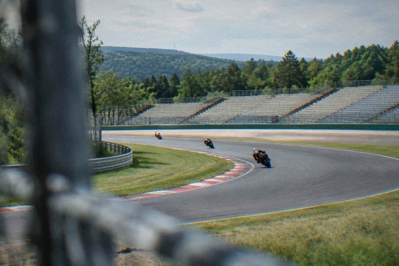 Motorcycle riders leaning into a fast corner on a wide racing circuit with grandstands in the background