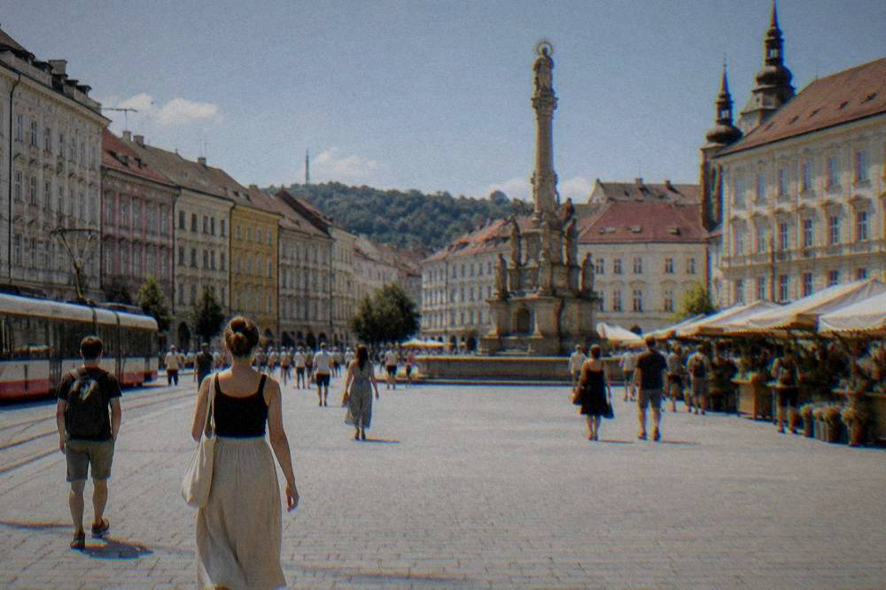 People walking across a wide cobbled city square with a baroque column and market stalls
