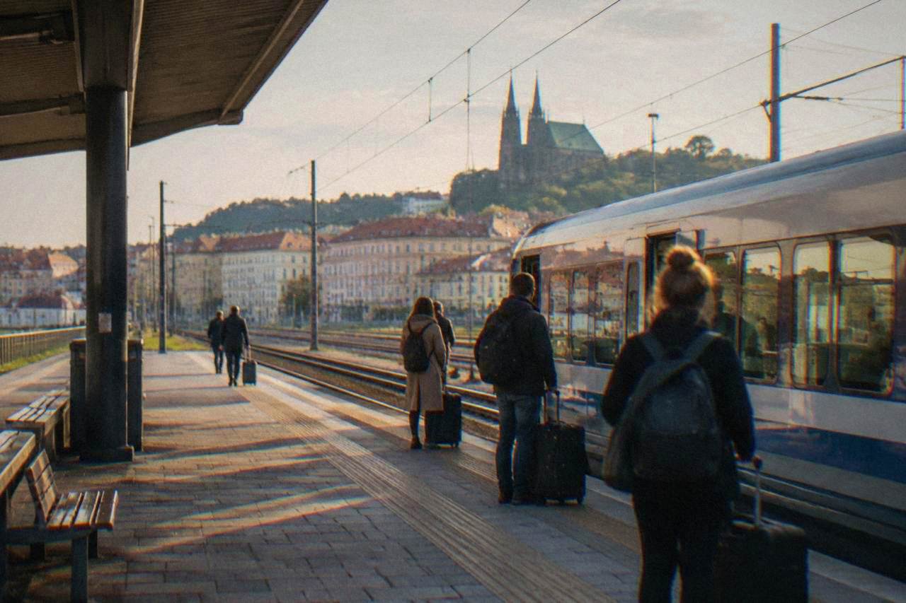 A railway station platform in Brno with a train at the platform and passengers waiting with luggage