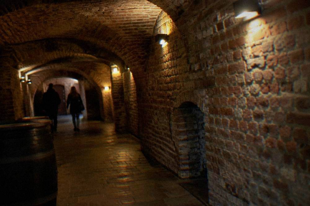Dimly lit underground brick cellar corridor with arched ceilings and warm lamp light