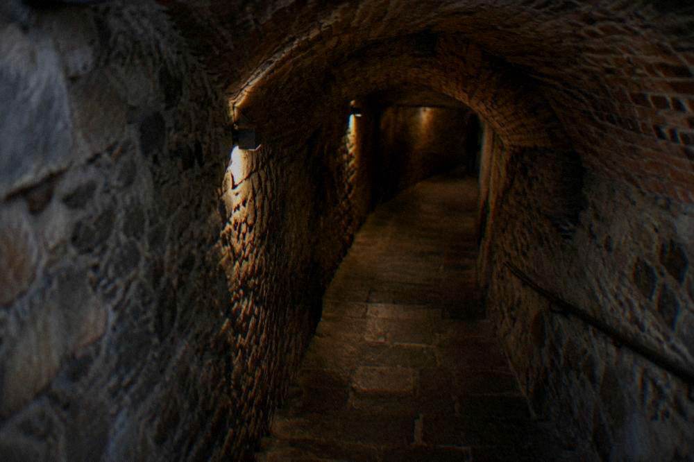 Narrow tunnel with stone walls and warm lamp lighting leading under a city square